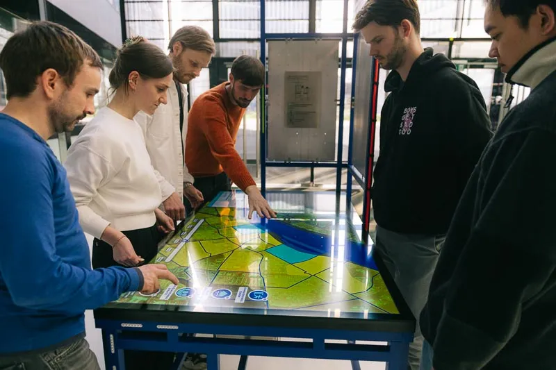 Sechs Personen stehen um einen interaktiven Tisch mit einer Landkarte in einer hellen Ausstellungshalle.