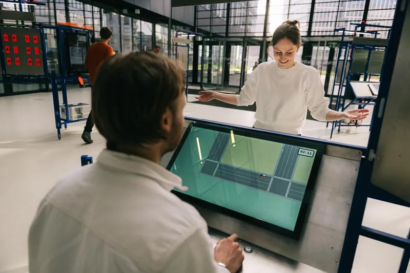 Zwei Personen spielen an gegenüberliegenden Seiten der interaktiven Station ein Spiel. Ein Mann bedient einen Touchscreen mit einem Stadtplan, während ihm gegenüber eine lächelnde Frau steht.