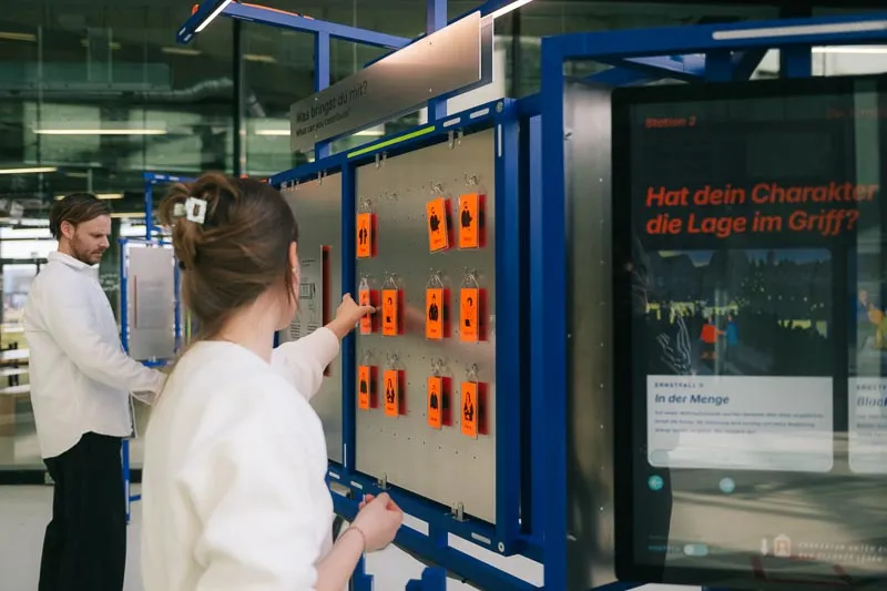Zwei Personen an der Station. Eine hängt orangefarbene Anhänger an eine Lochwand neben einem Touchscreen.