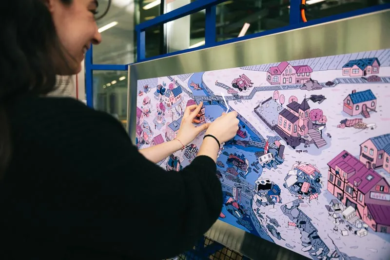 Eine Frau setzt ein Puzzleteil auf eine illustrierte Tafel mit dem Bild einer überfluteten Ortschaft.