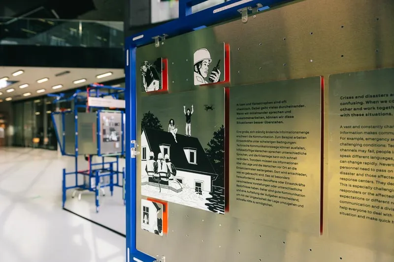 Nahaufnahme einer Ausstellungstafel mit Illustrationen im Comicstil und deutsch- und englischsprachigem Text auf einer Metallplatte.